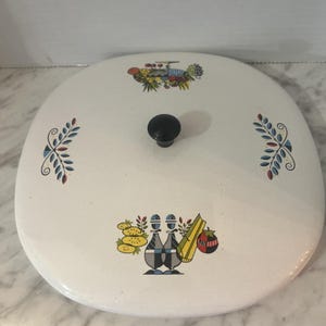 Può includere: Coperchio in smalto bianco con pomello nero e illustrazioni colorate di frutta e verdura. Il coperchio ha una forma quadrata arrotondata e motivi decorativi a foglie blu, rosse e gialle.