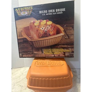 May include: A square terracotta-colored clay cooker with a matching lid featuring the embossed text OVEN BRIQUE. The product is displayed in front of its original cardboard packaging, which shows a 3 qt. capacity and a cooked ham inside the cooker.