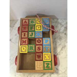 Puede incluir: Carro de bloques de alfabeto de madera con ruedas rojas y un asa de cuerda. El carro contiene bloques coloridos con las letras de la A a la Z. Los bloques son de varios colores, incluyendo rojo, verde, amarillo y azul. Un búho está tallado en un bloque.