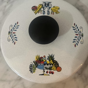 Op de afbeelding: Een witte emaille deksel met een zwarte knop. De deksel heeft kleurrijke illustraties van fruit, groenten en andere voedingsmiddelen. Er zijn ook decoratieve blad- en bloempatronen. De deksel is waarschijnlijk voor een pot of pan.