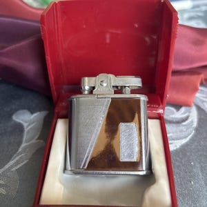 May include: A vintage chrome and brown lighter with a geometric design, presented in a red case. The lighter has a rectangular shape with a silver top and a brown panel. The case is open, revealing the lighter nestled inside.