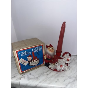 May include: A red metal candle holder featuring a small elf figure with a white beard and red hat, holding a red taper candle. The base is decorated with white pinecone-like ornaments. Beside it sits a cardboard box labelled White Christmas Pixie Candle Holder.
