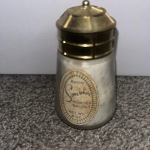 Op de afbeelding: Een vintage Avon Somewhere Powder Sachet fles. De fles is cilindrisch met een goudkleurig metalen deksel en basis. Het etiket luidt "Avon Somewhere Powder Sachet Net Wt. .9 oz."