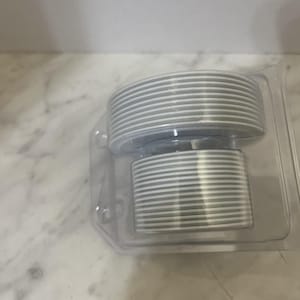 May include: A set of gray and white striped coasters, stacked in a clear plastic container. The coasters appear to be made of a hard material and are designed to protect surfaces from drink condensation. The container is rectangular with rounded corners.