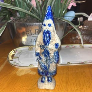Op de afbeelding: Een keramieken beeldje van een bebaarde kabouter met een puntmuts. De kabouter is voornamelijk beige met blauwe accenten op de muts, baard en kleding. Het beeldje heeft een textuur en staat op een kleine voet.