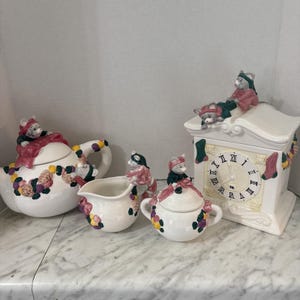 May include: A white ceramic tea set comprising a teapot, creamer, sugar bowl, and a clock-shaped canister. Each piece is adorned with sculpted mice wearing pink and green clothing, alongside colourful floral accents and hanging stocking motifs.