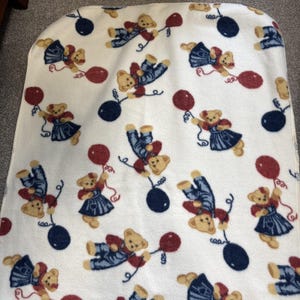 May include: A white fleece blanket with a repeating pattern of teddy bears holding red balloons and floating near navy blue balloons. The teddy bears are wearing blue denim overalls or dresses. The blanket has a rounded top edge.
