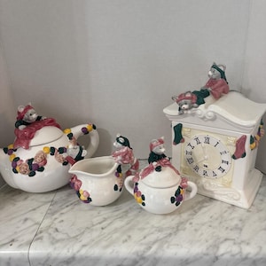 May include: A white ceramic tea set comprising a teapot, creamer, sugar bowl, and a clock-shaped canister. Each piece is adorned with sculpted mice wearing pink and green clothing, alongside colourful floral accents and hanging stocking motifs.
