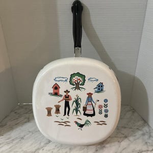 Könnte beinhalten: Weiß emaillierte Bratpfanne mit schwarzem Griff. Die Bratpfanne zeigt eine farbenfrohe Bauernhofszene mit einem Bauern, einer Frau, einem Hahn und verschiedenen Hofelementen. Der Griff ist etwa 25 cm lang.