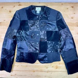 May include: A patchwork jacket in shades of navy blue, featuring a mix of textures including velvet and satin. The jacket has a round collar, button closure, and floral embellishments. The sleeves are long, and the jacket is displayed on a wooden surface.