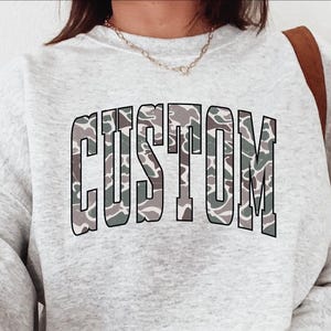 May include: A light gray sweatshirt with the word "CUSTOM" in large, block letters across the front. The letters have a camouflage pattern in shades of green, gray, and brown. The sweatshirt has long sleeves and a crew neck.