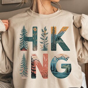 May include: A sand-colored crewneck sweatshirt featuring the word HIKING printed in large letters. Each letter is filled with nature-themed illustrations including evergreen trees, mountain peaks, a lake, and various plants in shades of blue, green, and peach.
