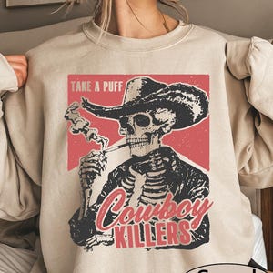 May include: Sand-colored sweatshirt featuring a graphic of a skeleton cowboy smoking a cigarette, wearing a hat and jacket. The design includes the text "TAKE A PUFF" and "Cowboy Killers" in red and white. The sweatshirt is a casual style.