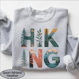 A gray sweatshirt with the word "HIKING" printed on it, featuring various nature-inspired illustrations such as pine trees, mountains, and forests.