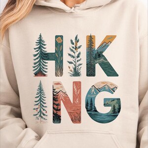 A woman wearing a beige hoodie with the word "HIKING" printed on it, featuring various nature-inspired illustrations such as pine trees, mountains, and a river.
