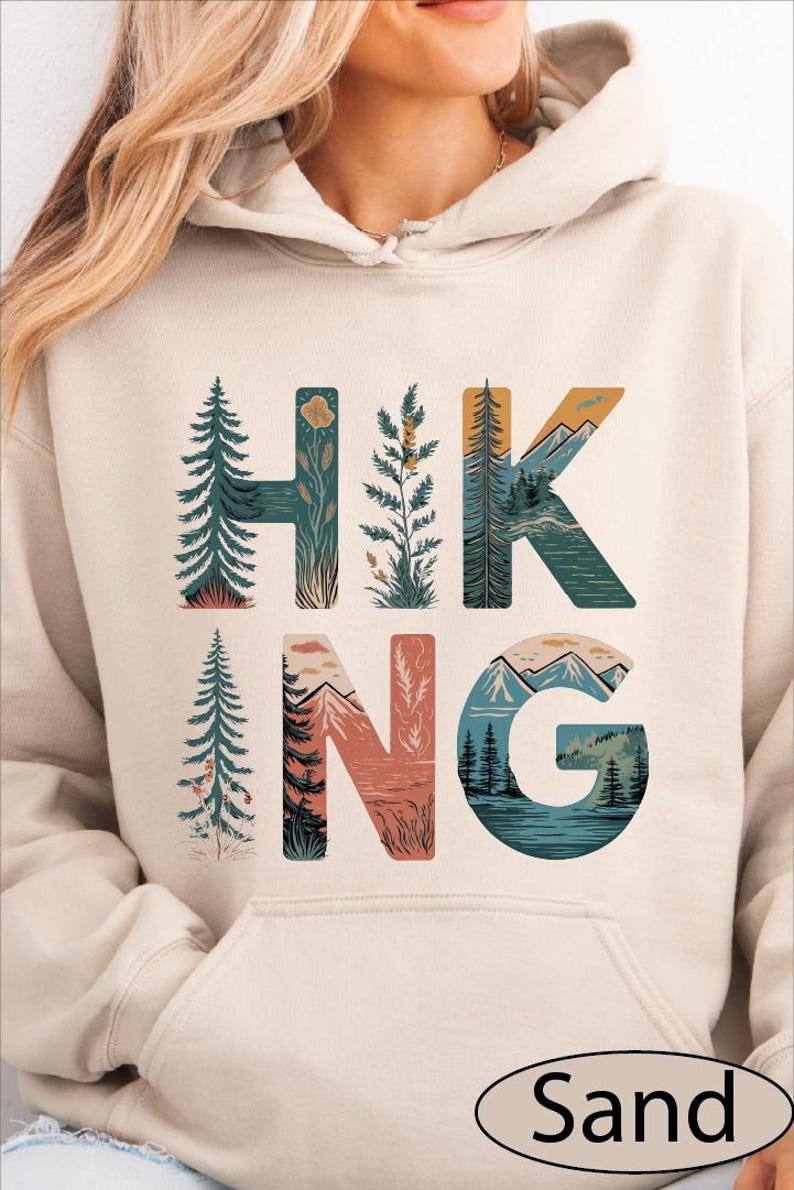 A woman wearing a beige hoodie with the word "HIKING" printed on it, featuring various nature-inspired illustrations such as pine trees, mountains, and a river.