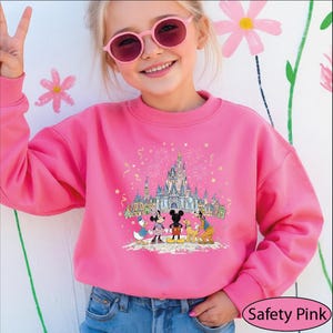 A young girl wearing a pink sweater with a Disney castle and characters on it, smiling and waving.