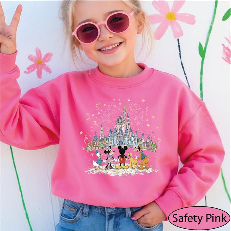 A young girl wearing a pink sweater with a Disney castle and characters on it, smiling and waving.