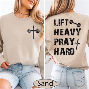 May include: A sand-colored crewneck sweatshirt featuring a black graphic of a barbell shaped like a cross on the front left chest. The back displays the black distressed text LIFT HEAVY PRAY HARD with a barbell and a cross icon integrated.