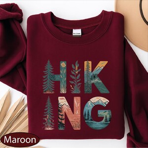 A maroon sweatshirt with the word "HIKING" printed on it, featuring various nature-inspired illustrations such as pine trees, mountains, and a river.