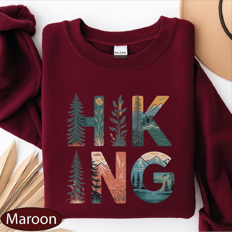 A maroon sweatshirt with the word "HIKING" printed on it, featuring various nature-inspired illustrations such as pine trees, mountains, and a river.