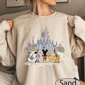 A woman wearing a beige sweater with a Disney-themed graphic of Minnie Mouse, Donald Duck, Goofy, and Mickey Mouse standing in front of a castle.