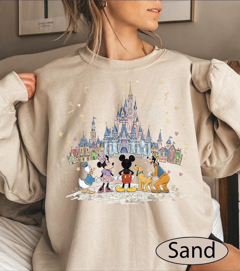 A woman wearing a beige sweater with a Disney-themed graphic of Minnie Mouse, Donald Duck, Goofy, and Mickey Mouse standing in front of a castle.