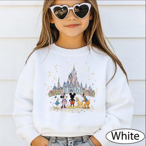 A young girl wearing a white sweater with a Disney castle and characters printed on it, smiling and posing for the camera.