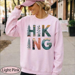 May include: A light pink long-sleeve sweatshirt featuring the word HIKING printed in large letters. Each letter is filled with a detailed nature illustration, including evergreen trees, mountain ranges, and a lake scene in shades of green, blue, and orange.