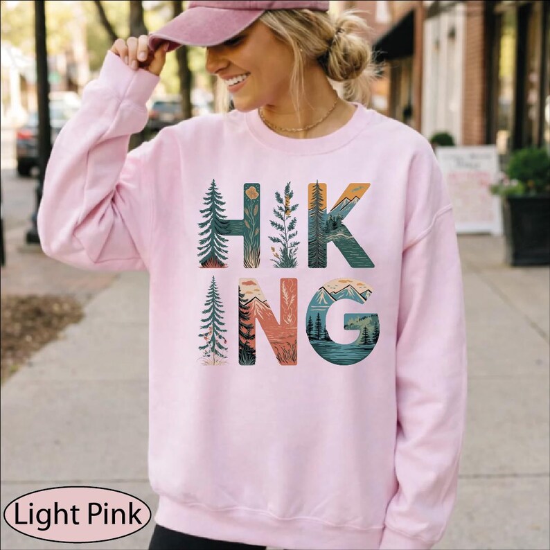 May include: A light pink long-sleeve sweatshirt featuring the word HIKING printed in large letters. Each letter is filled with a detailed nature illustration, including evergreen trees, mountain ranges, and a lake scene in shades of green, blue, and orange.