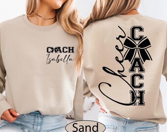 May include: Sand-colored sweatshirt with the text "COACH Isabella" on the front and a black bow graphic. The back features the word "Cheer" in script and the word "COACH" in block letters with a bow graphic.