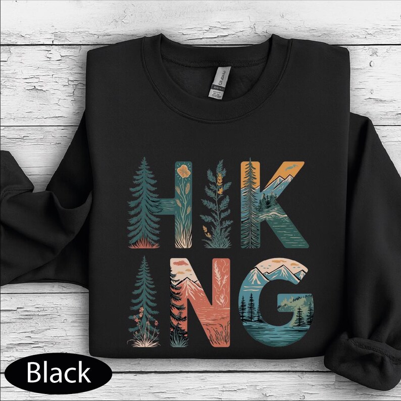 A black t-shirt with a graphic design that says "HIKING" in a stylized font, featuring various elements such as trees, mountains, and flowers.