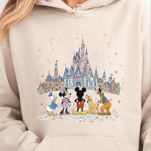 A woman wearing a beige hoodie with a Disney-themed graphic of Mickey Mouse, Minnie Mouse, Donald Duck, Goofy, and other Disney characters standing in front of a castle.