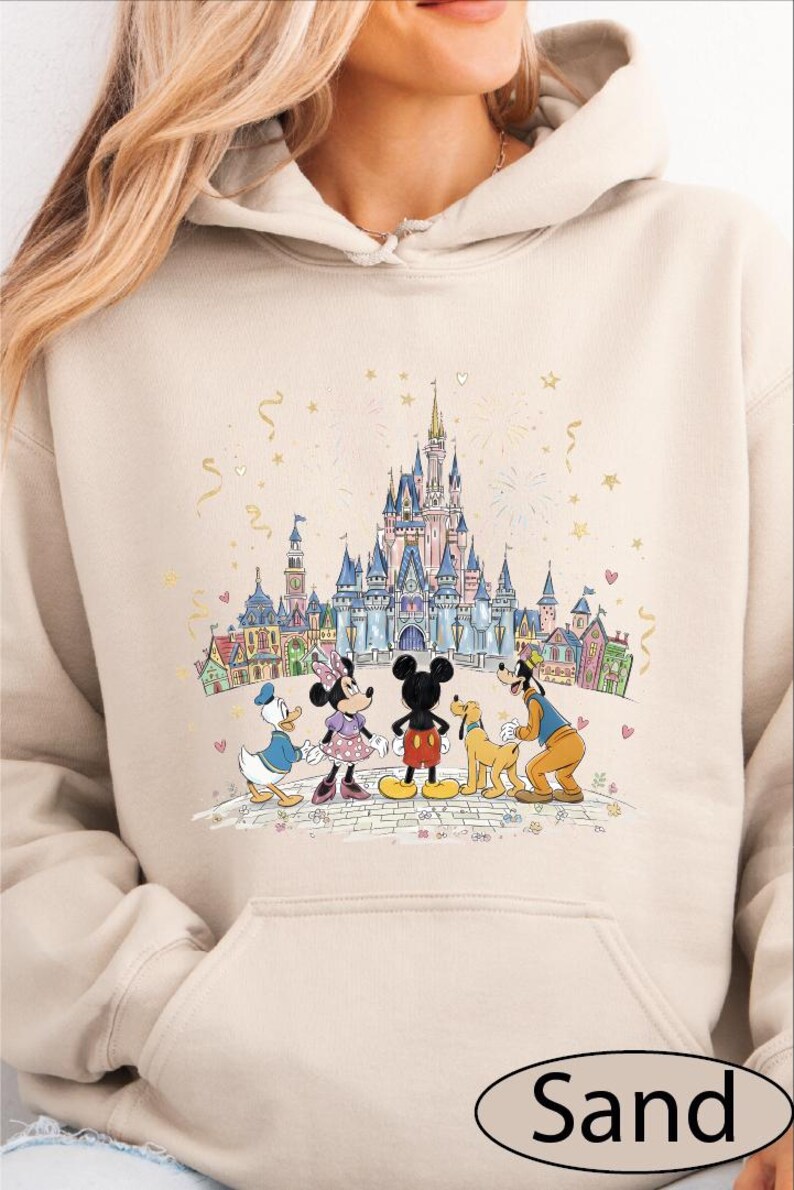 A woman wearing a beige hoodie with a Disney-themed graphic of Mickey Mouse, Minnie Mouse, Donald Duck, Goofy, and other Disney characters standing in front of a castle.