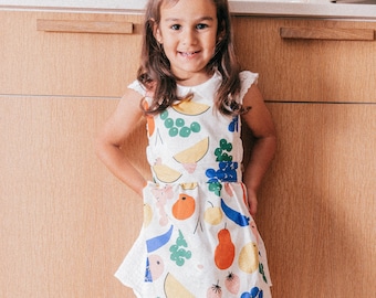 Puede incluir: Una niña pequeña con un vestido blanco con estampado de frutas de colores. Está de pie frente a un armario de madera y lleva un lazo blanco en el pelo.