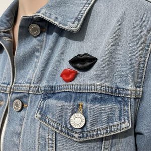 May include: A light blue denim jacket featuring two lip-shaped pins attached to the chest area. One pin is a larger black set of lips, and the other is a smaller, bright red set of lips. The jacket has silver-toned metal buttons and stitching.
