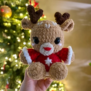 Peut inclure: Une peluche de renne au crochet faite à la main. Le renne a un corps beige, des bois bruns et de grands yeux noirs. Il porte un manteau rouge avec une bordure blanche et une étoile blanche sur le devant. Le jouet est conçu pour la période des fêtes.