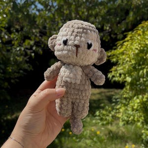 Op de afbeelding: Een handgemaakte gehaakte apenpop in beige en bruin. De pop heeft een getextureerd, fuzzy uiterlijk met kleine oren, een knoopneus en zwarte ogen. De aap wordt vastgehouden tegen een vage groene achtergrond.