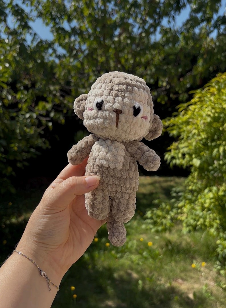 Op de afbeelding: Een handgemaakte gehaakte apenpop in beige en bruin. De pop heeft een getextureerd, fuzzy uiterlijk met kleine oren, een knoopneus en zwarte ogen. De aap wordt vastgehouden tegen een vage groene achtergrond.
