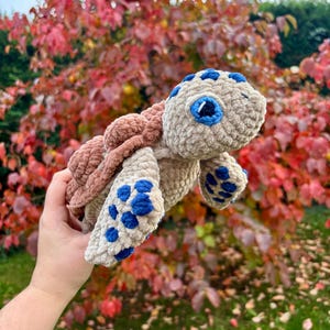 Peut inclure: Une tortue en peluche crochetée, beige, marron et bleu. La tortue a une carapace texturée, des nageoires à pois bleus et des yeux bordés de bleu. Le jouet en peluche est conçu pour ressembler à une tortue de mer.