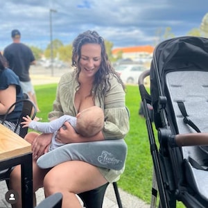 Könnte beinhalten: Eine Frau stillt ein Baby, während sie auf einem Stuhl im Freien sitzt. Sie verwendet ein graues Stillkissen mit dem Aufdruck "Feeding Support". Im Hintergrund steht ein Kinderwagen.