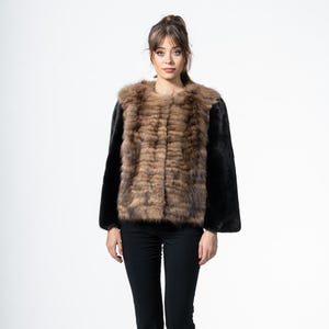 Può includere: Una giacca di pelliccia marrone e nera con maniche nere. La giacca è indossata con pantaloni neri e scarpe nere. La modella è in piedi davanti a uno sfondo bianco.