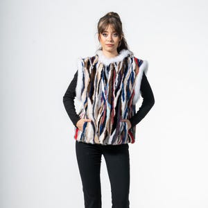 May include: A multi-coloured fur vest with a white fur trim. The vest features vertical stripes in shades of brown, blue, red, and white. The model is wearing a black long-sleeved shirt and black trousers.
