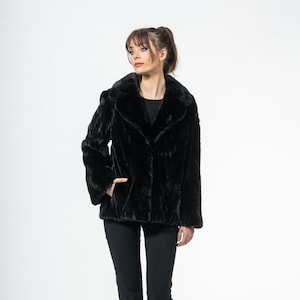 Puede incluir: Chaqueta de piel sintética negra con cuello ancho y mangas largas. La chaqueta se lleva con pantalones negros y zapatos negros. La modelo está de pie frente a un fondo blanco.