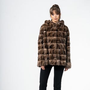 Könnte beinhalten: Braune Kapuzenjacke aus Fell mit geometrischem Muster. Die Jacke wird mit schwarzer Hose und schwarzen Absätzen getragen. Das Fell scheint eine Mischung aus hellen und dunklen Brauntönen zu sein. Der Hintergrund ist schlicht weiß.