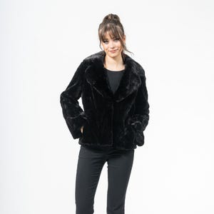 Può includere: Giacca nera in ecopelliccia con colletto classico e chiusura con bottoni. La giacca è abbinata a pantaloni neri e tacchi neri. L'outfit è ambientato su uno sfondo bianco, creando un look pulito e moderno.