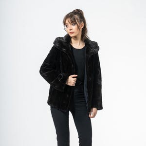 May include: A black faux fur jacket with a hood. The jacket is buttoned and worn with black pants and black heels. The model is standing in front of a white background.