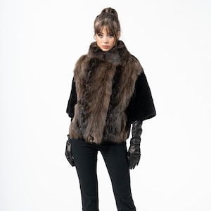 Può includere: Una donna indossa un gilet di pelliccia marrone con maniche nere, pantaloni neri e guanti di pelle neri. Il gilet ha un colletto alto e un aspetto strutturato. L'outfit è completato da tacchi neri.