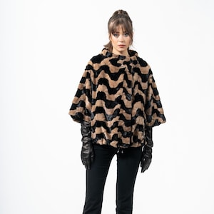 May include: A faux fur cape in a brown and black zigzag pattern. The cape has a high collar and wide sleeves. The model is wearing black gloves, black pants, and black heels. The background is white.