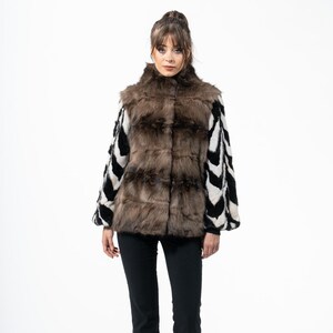 May include: A woman wearing a brown and black fur vest with black and white patterned sleeves, paired with black pants and shoes. The fur vest has a high collar and a textured appearance. The sleeves feature a bold chevron design.